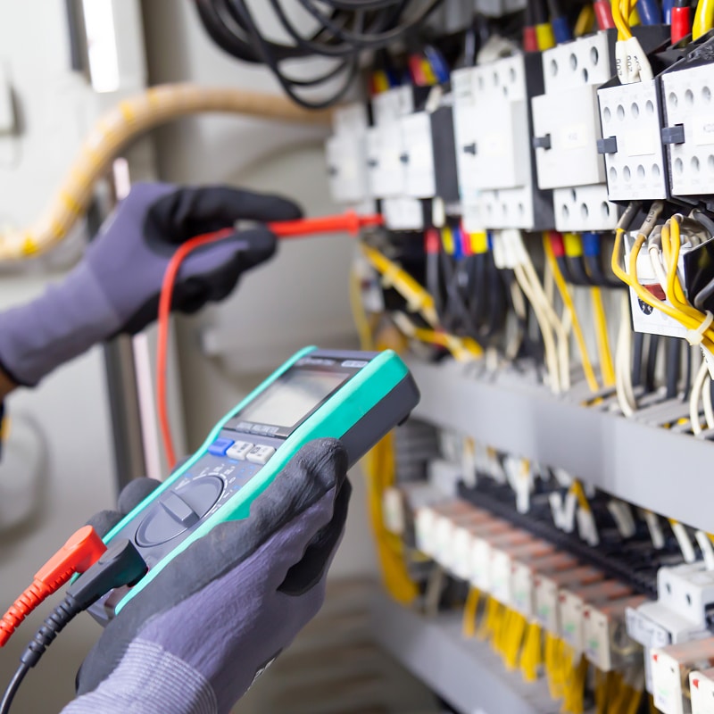 Électricien ganté mesurant des contacteurs dans un tableau électrique avec un multimètre numérique teal et des sondes rouges.