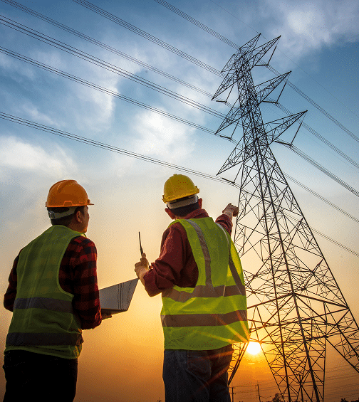 Ingénieurs électriciens : inspection de pylône et réseau Deux ingénieurs en casques et gilets de sécurité observent un pylône électrique géant et ses lignes de transmission au coucher du soleil.