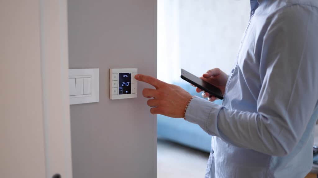 Contrôlez votre maison avec un thermostat intelligent connecté Main d'homme ajustant un thermostat intelligent affichant 24.0°C et 10:46 (SUN, SYSTEM, Man), tout en tenant un smartphone. Gestion domotique.